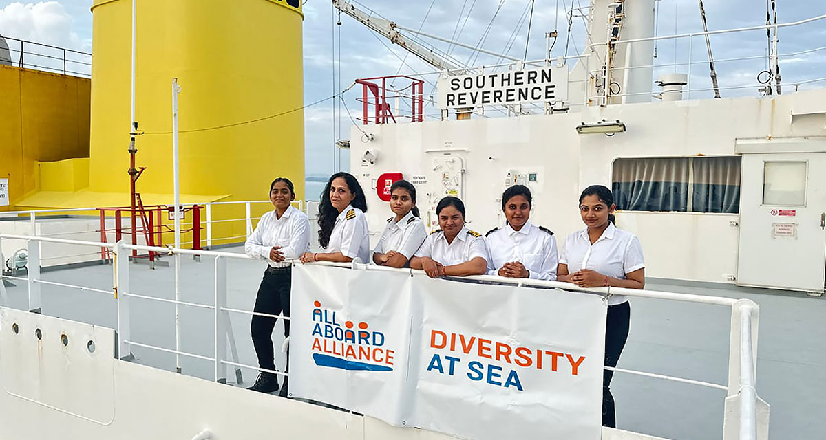 Six women seafarers in uniform standing on the deck of the Southern Reverence ship, next to a banner that reads ‘Diversity at Sea’ and ‘All Aboard Alliance.