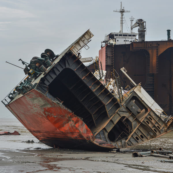 Partially dismantled ship resting on the muddy shore at a ship recycling yard, with a rusted hull and tilted structure