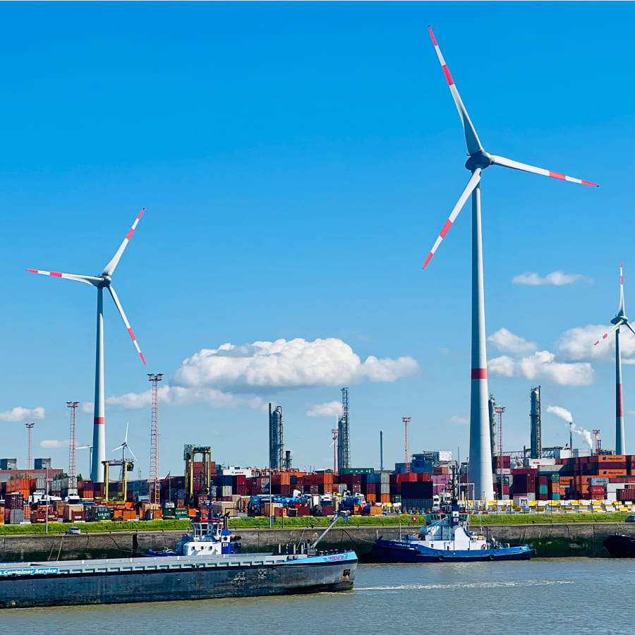 Wind turbines stand tall near a busy port filled with containers and docked vessels, under a bright blue sky