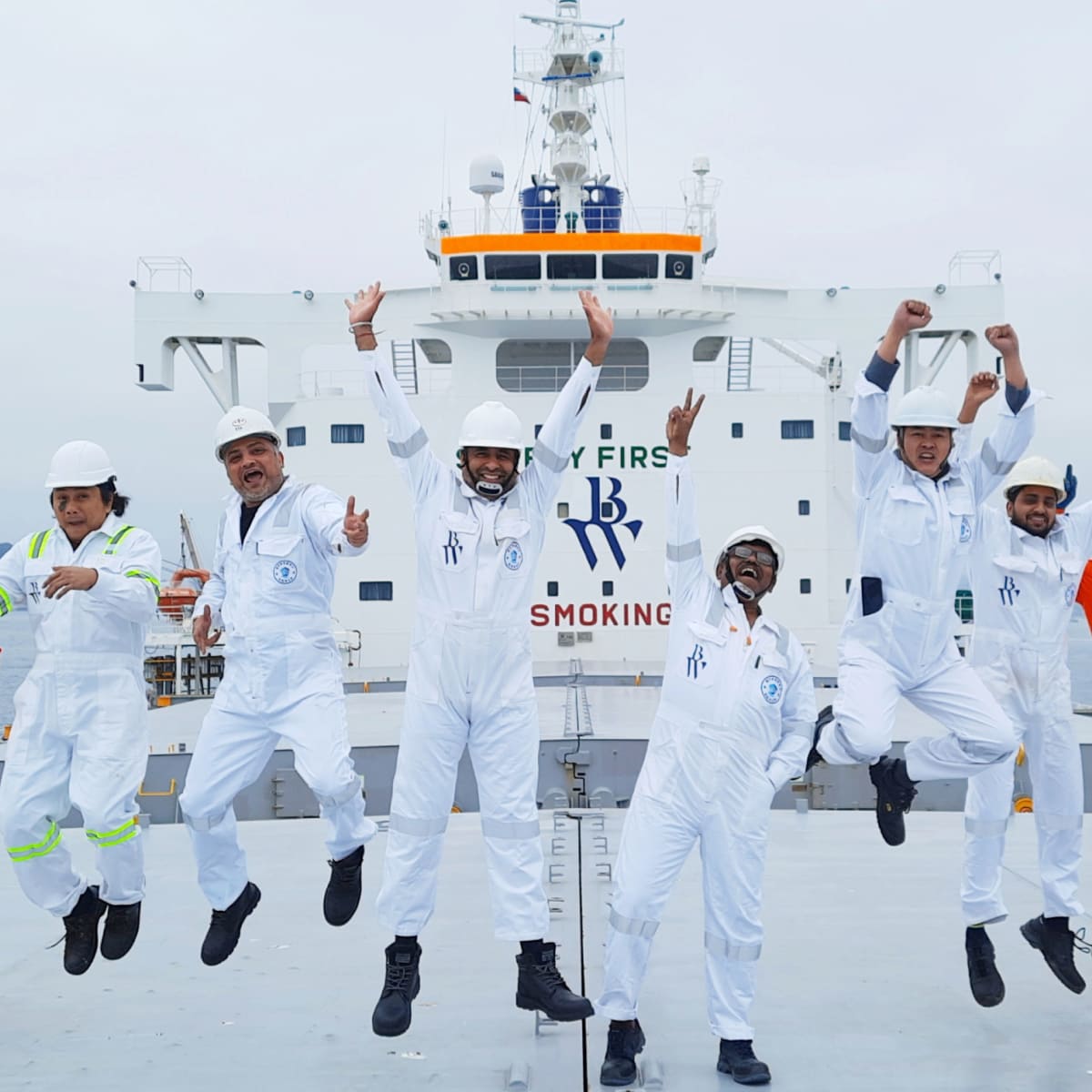 Group of seafarers in white coveralls and helmets jumping joyfully on a ship’s deck, with the words “Safety First” and “No Smoking” visible on the ship’s bridge