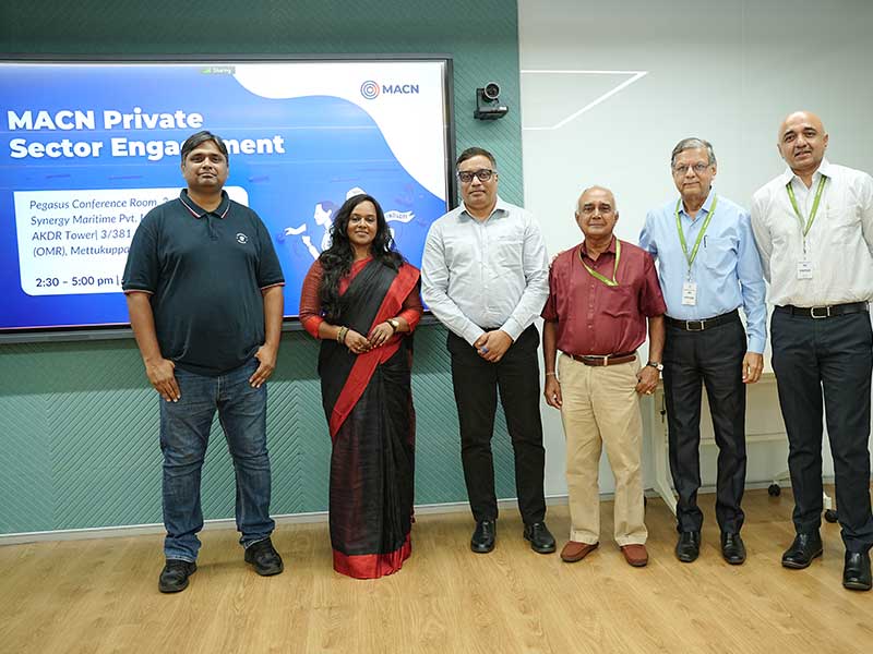 Group of six participants at the MACN event posing together in front of a presentation screen, dressed in formal business attire