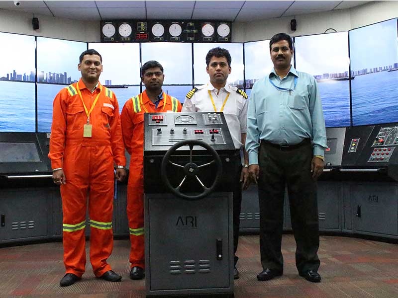 Four maritime professionals in uniform posing in a ship simulation room, standing behind a training helm labeled “ARI.”