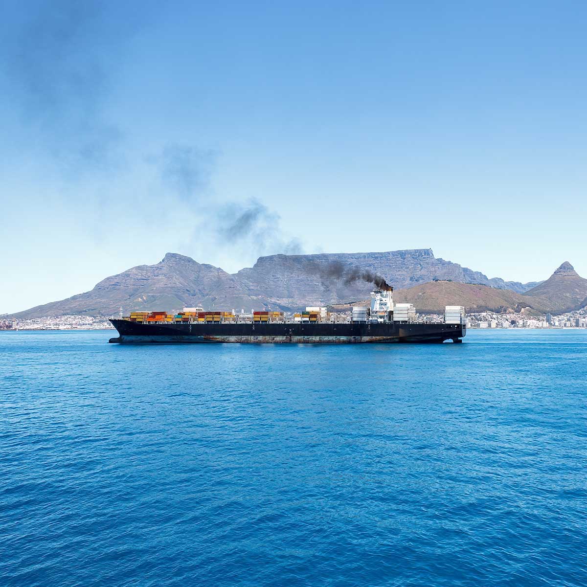Cargo ship sailing near a city coastline with mountains in the background under a clear sky