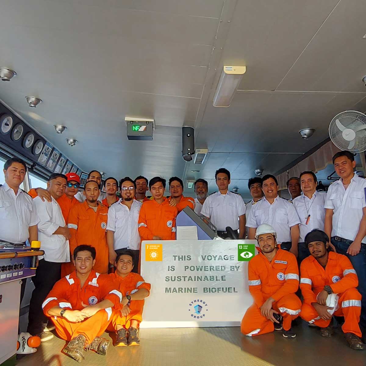 Group of crew members in orange and white uniforms posing inside a ship’s control room beside a sign that reads “This voyage is powered by sustainable marine biofuel.”