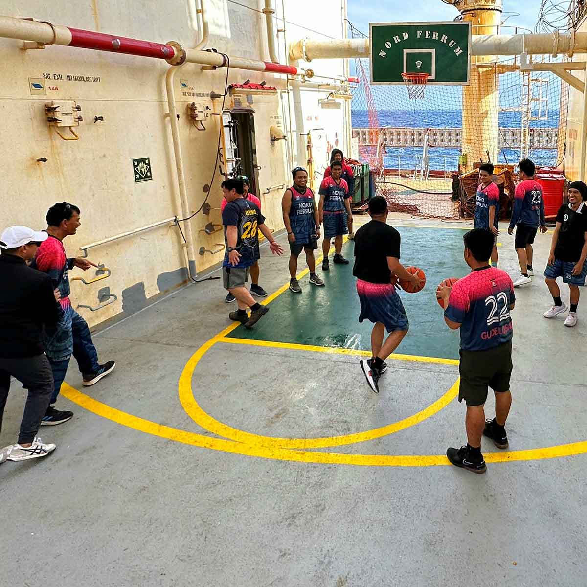 Crew members playing basketball on a ship’s deck, wearing matching jerseys with “Nord Ferrum” printed on them, with the ocean visible in the background