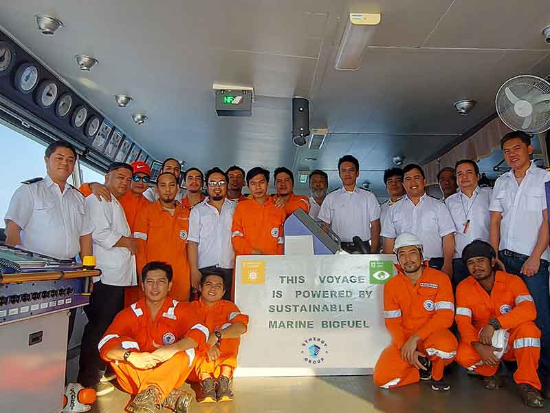 Crew members wearing orange coveralls and white uniforms pose on a ship’s bridge around a sign reading “This Voyage Is Powered By Sustainable Marine Biofuel,” promoting renewable energy use at sea